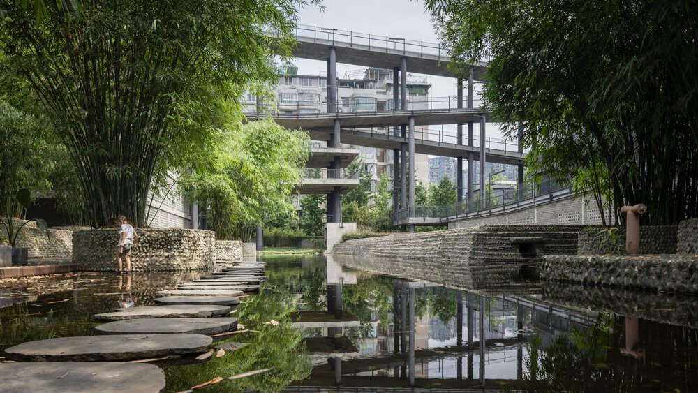 Eine Person steht an einem Teich mit Steinstufen, umgeben von Bambus und moderner Architektur im Hintergrund.