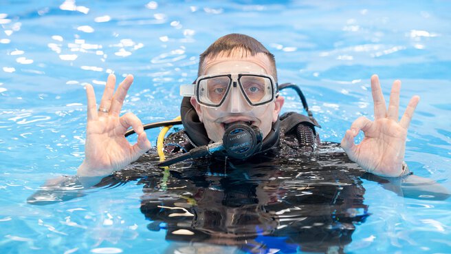 A person wearing diving equipment is standing in a pool and making the “okay” sign with both hands. The person is wearing a diving mask and a regulator, and the water around them is calm.