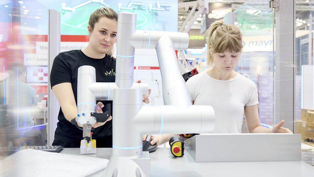 Two women operate a robotic arm at a technical exhibition stand with various devices around them.