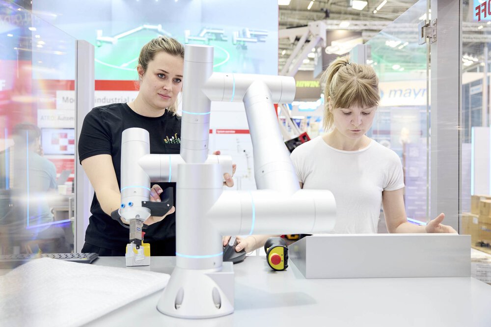 Zwei Frauen bedienen einen Roboterarm an einem technischen Messestand mit verschiedenen Geräten um sie herum.