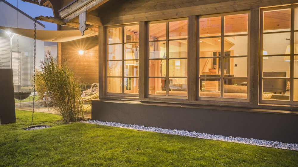 Gemütliches Holzhaus mit großen, von innen beleuchteten Fenstern, umgeben von einem üppigen grünen Rasen in der Abenddämmerung.