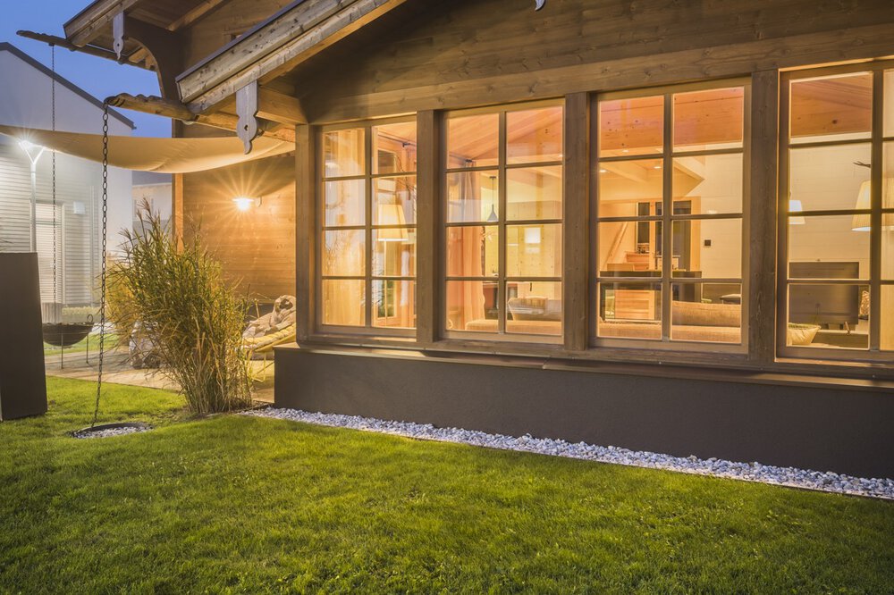 Gemütliches Holzhaus mit großen, von innen beleuchteten Fenstern, umgeben von einem üppigen grünen Rasen in der Abenddämmerung.
