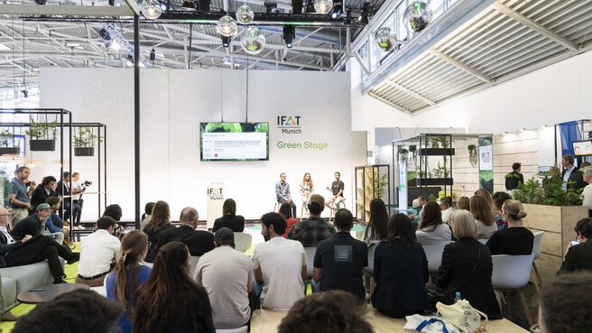 Audience seated in front of the IFAT Munich Green Stage, where three people are sitting on stools and speaking.