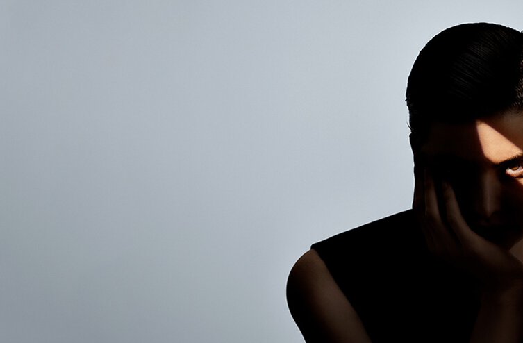 Woman holding a blue gemstone to her eye, with dramatic shadows on her face against a gray background.