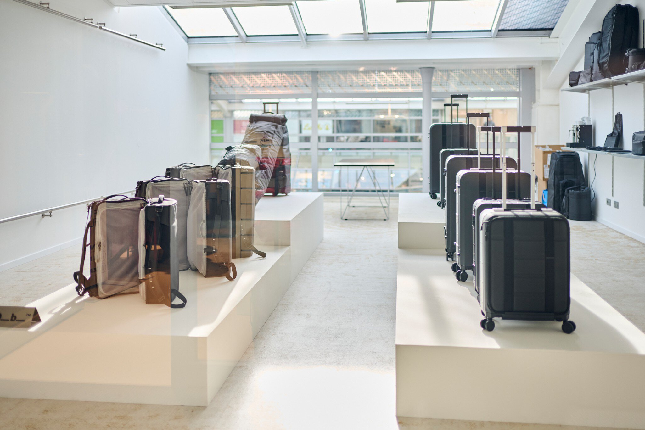 Ein weißer Ausstellungsraum mit Oberlichtern und einer Glaswand. Davor stehen auf Podesten diverse Rollkoffer und Reisetaschen zur Ansicht.