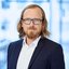 Man with glasses and long dark blond hair in a suit with white shirt.