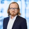 Man with glasses and long dark blond hair in a suit with white shirt.