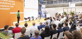 Ein humanoider Roboter steht auf einer Bühne mit mehreren Sprechern, während ein großes Publikum auf einer Messe zuhört.