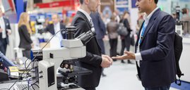 Two businessmen are talking, in the foreground there is a microscope, in the background an exhibition hall with other stands can be seen.