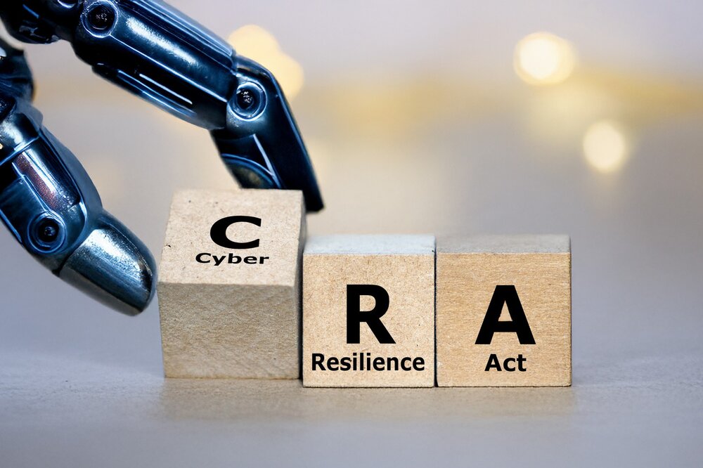 Eine detaillierte Nahaufnahme einer mechanischen, metallisch glänzenden Roboterhand, die drei nebeneinander liegende Holzwürfel berührt. Auf den Würfeln sind die Buchstaben „C“, „R“ und „A“ aufgedruckt, jeweils ergänzt durch die Wörter „Cyber“, „Resilience“ und „Act“. Der Hintergrund ist in warmen, unscharfen Lichttönen (Bokeh-Effekt) gehalten.