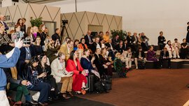 The audience sits in rows and attentively follows an event in a modern hall with geometric décor. Jewellery artists can be seen.