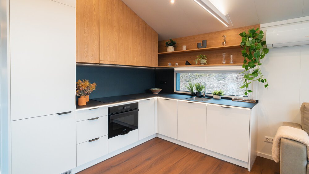 Eine moderne Küche in „L“-Form mit großen weißen Fronten, hölzernen Oberschränken, einem Herd, Regalbrettern und einem schmalen Fenster.