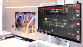 Nahansicht eines Computerbildschirms mit Tastatur, der an eine Maschine mit Fenster angeschlossen ist. Man erkennt bunte Kabel in der Maschine.