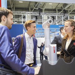 Drei Personen unterhalten sich an einem Messestand, zwischen Ihnen steht ein ausgestellter Roboterarm.