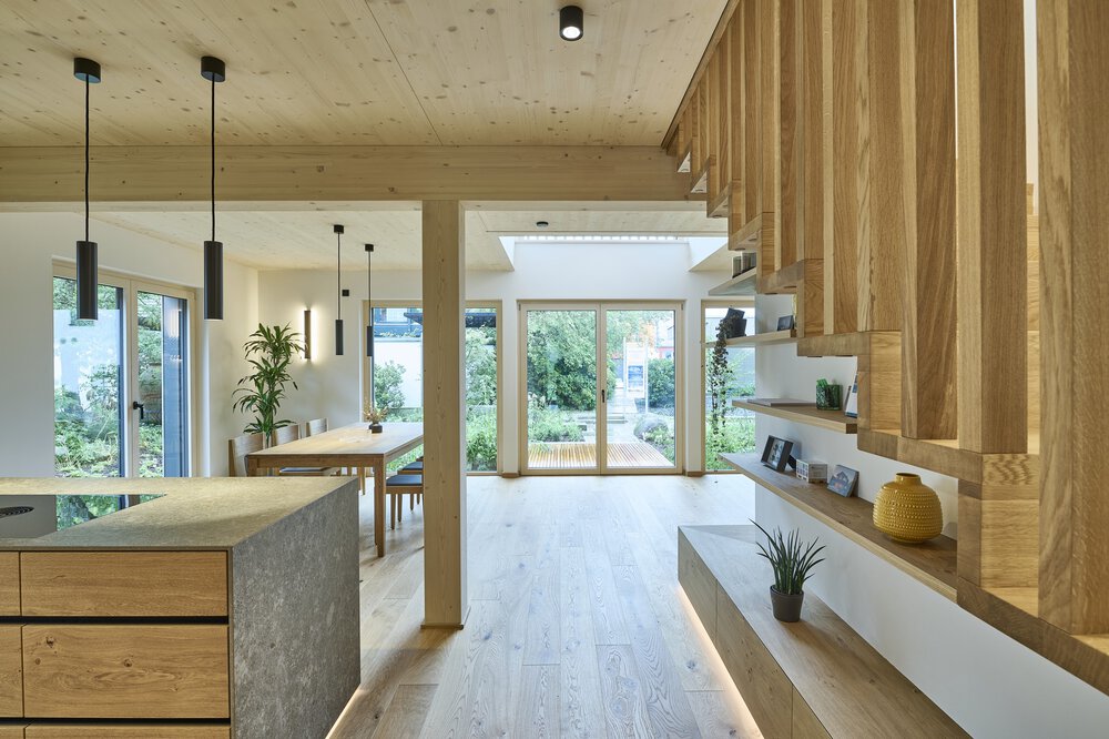 Moderne Küche mit Holzschränken, Hängelampen und großen Fenstern mit Blick ins Grüne.
