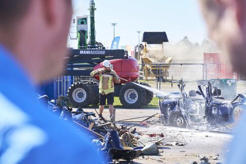 Ein Arbeiter auf einem Freigelände bearbeitet mit einem Hochdruckstrahl das Wrack eines Autos. Im Vordergrund sind unscharf ein paar Zuschauer zu sehen.