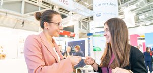Two women stand at a table in the careers area of electronica, smiling and exchanging business cards.