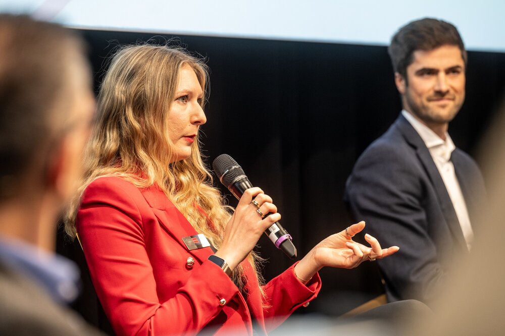 Eine Frau im roten Blazer spricht während einer Podiumsdiskussion in ein Mikrofon.