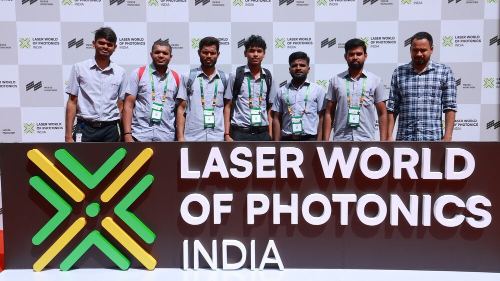 Seven men stand between the Laser World of Photonics India logo in front of them and a photo wall behind them, posing for a photo.