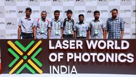 Seven men stand between the Laser World of Photonics India logo in front of them and a photo wall behind them, posing for a photo.