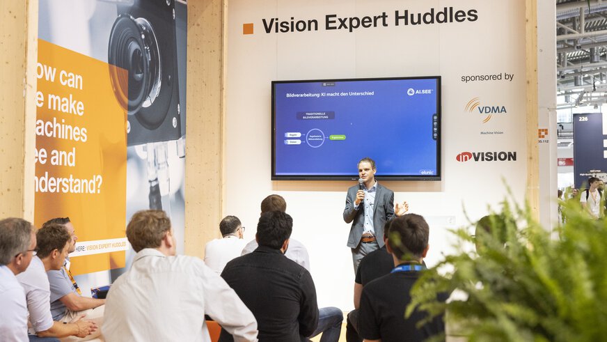 Ein Redner hält auf der Messe einen Vortrag zum Thema Bildverarbeitung mit Künstlicher Intelligenz im Rahmen der „Vision Expert Huddles“. Aufmerksame Zuhörer sitzen im Publikum, während auf einem großen Bildschirm eine Präsentationsfolie mit dem Titel „Bildverarbeitung: KI macht den Unterschied“ gezeigt wird. Logos von VDMA und inVISION sind sichtbar.