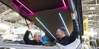 A woman and a man look out of an open car roof tent, which they are holding open with their arms, laughing. The car roof tent is in a large exhibition hall.