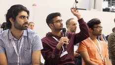 Drei Männer sitzen im Publikum, einer hält ein Mikrofon und hebt die Hand, um eine Frage zu stellen.