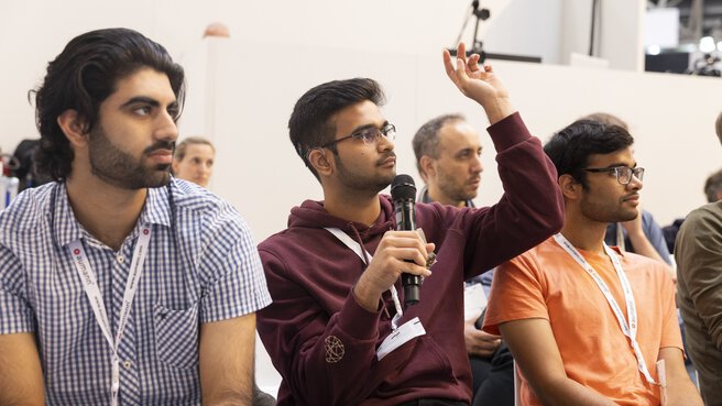 Three men sit in the audience, one holds a microphone and raises his hand to ask a question.