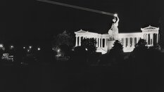 Schwarzweißfoto der Bavaria Statue bei Nacht, der Loorbeerkranz in der Hand der Statue wird von einem Laser angestrahlt und ist hell erleuchtet.