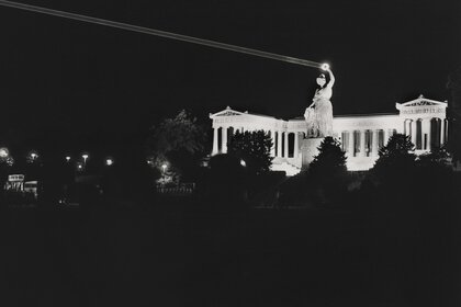 Schwarzweißfoto der Bavaria Statue bei Nacht, der Loorbeerkranz in der Hand der Statue wird von einem Laser angestrahlt und ist hell erleuchtet.
