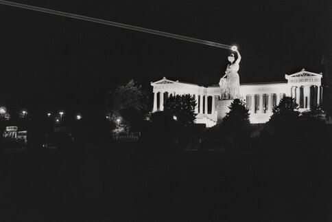 Black and white photo of the Bavaria statue at night, the laurel wreath in the statue's hand is illuminated by a laser and is brightly lit.