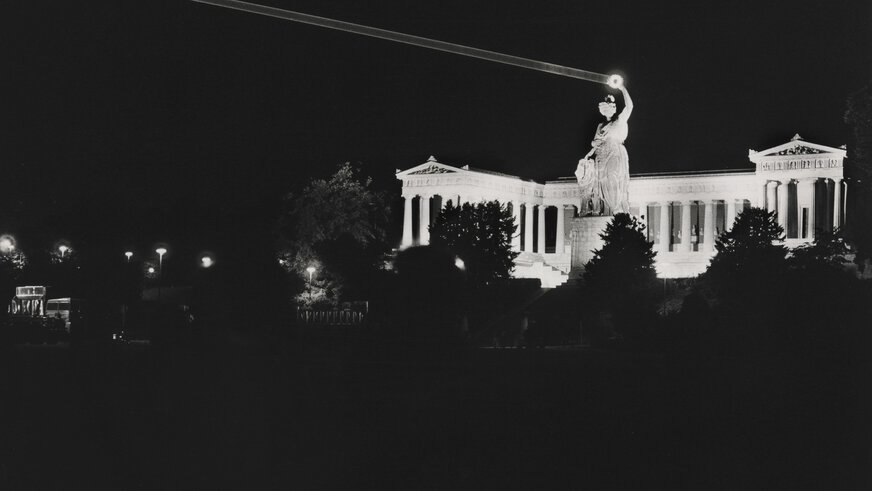 Schwarzweißfoto der Bavaria Statue bei Nacht, der Loorbeerkranz in der Hand der Statue wird von einem Laser angestrahlt und ist hell erleuchtet.