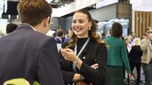 Two people are talking in front of an exhibition stand, one with her back to the camera. Other exhibition stands and visitors can be seen in the background.