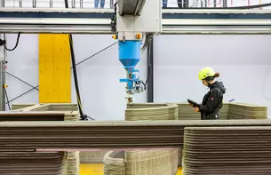 Eine Person in Sicherheitskleidung überwacht einen 3D-Drucker, der eine Struktur aus Beton fertigt.