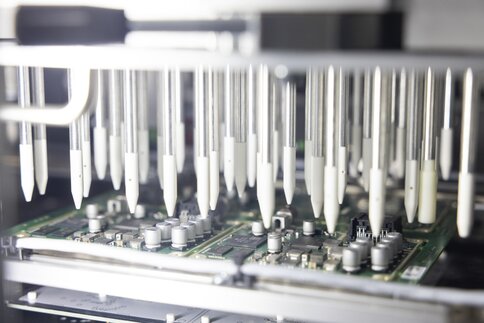 Close-up of a circuit board with electronic components, under a machine with many white rubber tips.