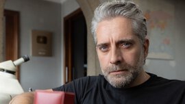 A man with short grey hair and a beard sits at a wooden table holding an open red jewellery box in his hands.