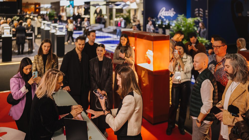 Menschen versammeln sich in einer belebten Ausstellungshalle um eine Schmuckausstellung und schauen sich eine Präsentation an.