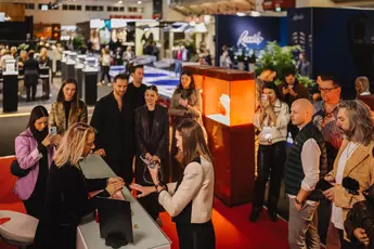 People gather around a jewelry exhibition in a busy exhibition hall and look at a presentation.