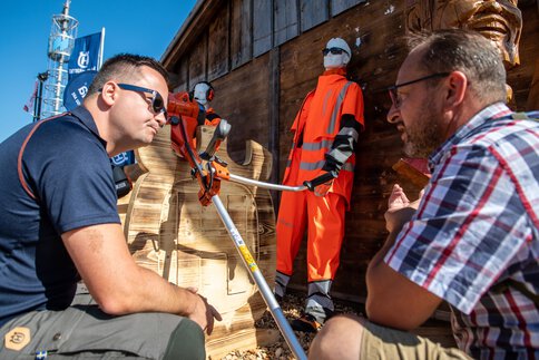  Zwei Männer betrachten eine Motorsäge an einem Messestand im Außenbereich, im Hintergrund Schutzkleidung auf Schaufensterpuppen.