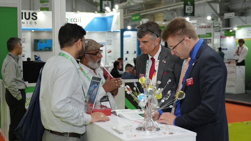 Four men are standing around a counter at a trade fair booth, talking to each other. There are colorful decorations on the counter.