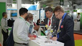 Four men are standing around a counter at a trade fair booth, talking to each other. There are colorful decorations on the counter.