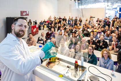 A man in a lab coat holds up a green liquid. In the background, many people are watching him experiment