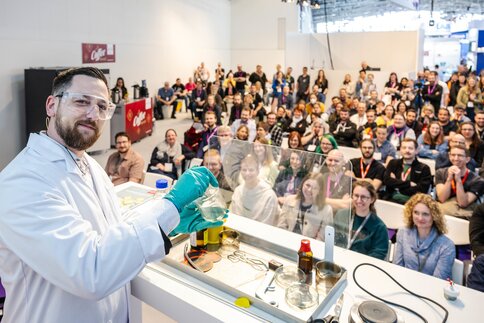 A man in a lab coat holds up a green liquid. In the background, many people are watching him experiment