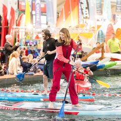Eine lachende Frau in einem roten Ganzkörperanzug steht barfuß auf einem Board im Pool der f.re.e Messe und paddelt. Hinter ihr weitere Messebesucher im und am Wasser.