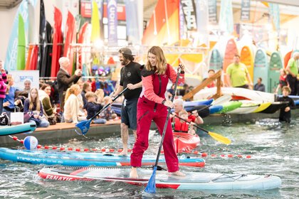 Eine lachende Frau in einem roten Ganzkörperanzug steht barfuß auf einem Board im Pool der f.re.e Messe und paddelt. Hinter ihr weitere Messebesucher im und am Wasser.