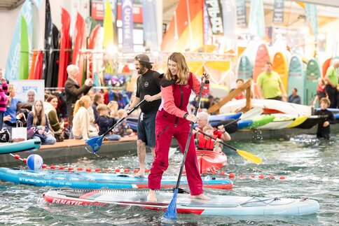 Eine lachende Frau in einem roten Ganzkörperanzug steht barfuß auf einem Board im Pool der f.re.e Messe und paddelt. Hinter ihr weitere Messebesucher im und am Wasser.