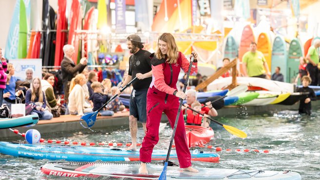 A smiling woman in a red full-body suit stands barefoot on a board in the pool at the f.re.e trade fair and paddles. Behind her are other trade fair visitors in and around the water.