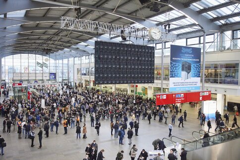 Blick über die Eingangshalle der productronica. Zahlreiche Besucher und gehen durch die Einlasskontrolle oder stehen in Grüppchen zusammen.