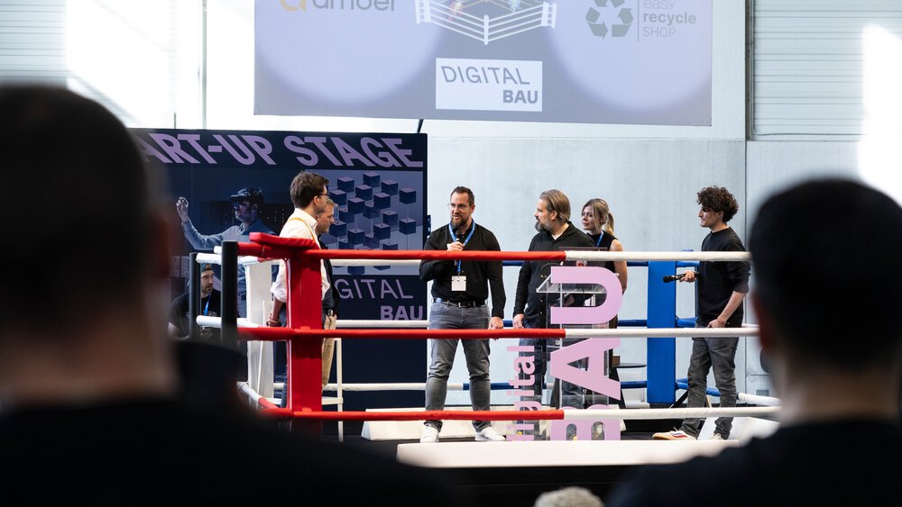 Fünf Personen stehen in einem Boxring auf einer Bühne mit der Aufschrift START-UP STAGE auf einer Tech-Veranstaltung, und das Publikum schaut zu.