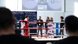 Fünf Personen stehen in einem Boxring auf einer Bühne mit der Aufschrift START-UP STAGE auf einer Tech-Veranstaltung, und das Publikum schaut zu.